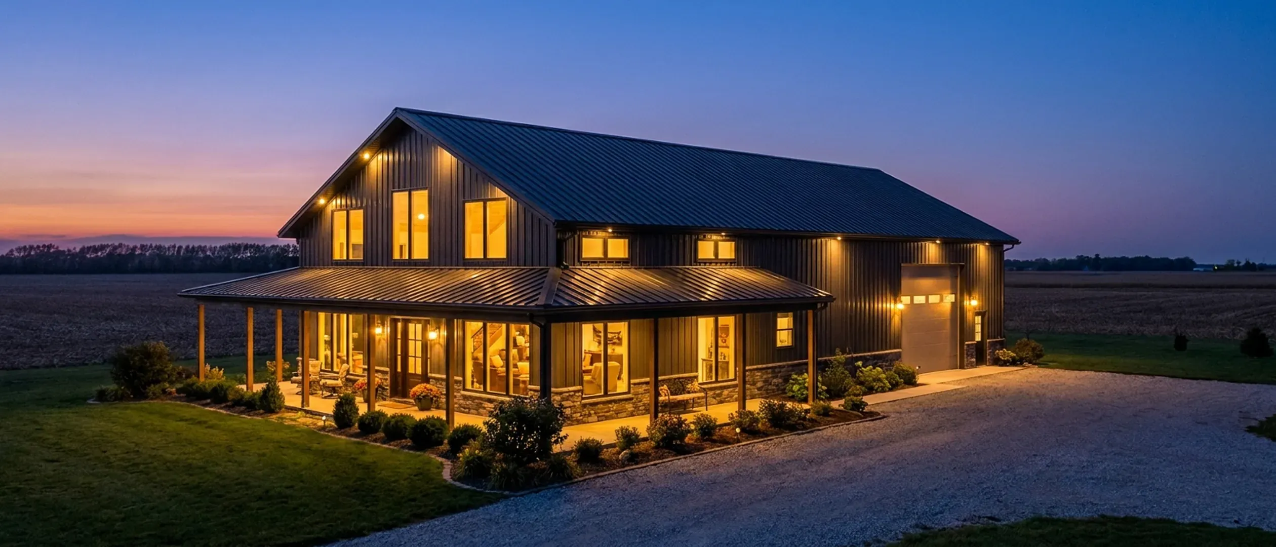 A barndominium at dusk with warm light glowing from every window and exterior lighting along the porch and roofline, flat farmland fading into a deep blue twilight sky behind it.